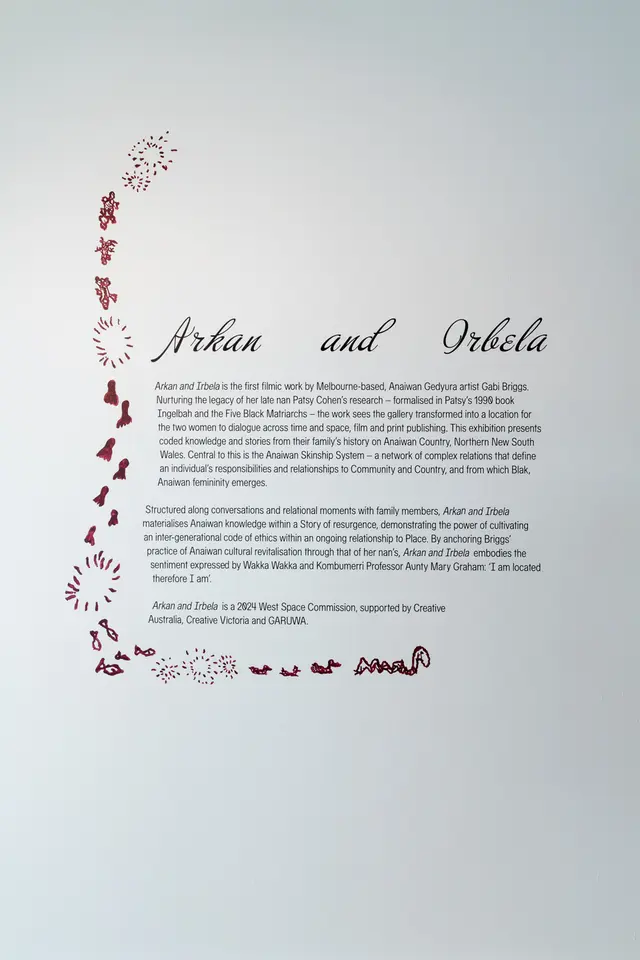 A wall has a paragraph of text titled Arkan and Irbela in cursive text with a cascading wall drawing in maroon depicting turtles, fish, a snake, footprints and markings. The text reads: "Arkan and Irbela is the first filmic work by Melbourne-based, Anaiwan Gedyura artist Gabi Briggs. Nurturing the legacy of her late nan Patsy Cohen's research-formalised in Patsy's 1990 book Ingelbah and the Five Black Matriarchs - the work sees the gallery transformed into a location for the two women to dialogue across time and space, film and print publishing. This exhibition presents coded knowledge and stories from their family's history on Anaiwan Country, Northern New South Wales. Central to this is the Anaiwan Skinship System - a network of complex relations that define an individual's responsibilities and relationships to Community and Country, and from which Blak, Anaiwan femininity emerges. Structured along conversations and relational moments with family members, Arkan and Irbela materialises Anaiwan knowledge within a Story of resurgence, demonstrating the power of cultivating an inter-generational code of ethics within an ongoing relationship to Place. By anchoring Briggs' practice of Anaiwan cultural revitalisation through that of her nan's, Arkan and Irbela embodies the sentiment expressed by Wakka Wakka and Kombumerri Professor Aunty Mary Graham: I am located therefore I am'.  Arkan and Irbela is a 2024 West Space Commission, supported by Creative Australia, Creative Victoria and GARUWA."