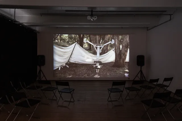 A projected image on the wall shows a long stretch of white fabric supported by a black pole. The fabric is stretched loosely across a natural environment of trees and dirt. A metal support structure is in the centre of the still. There are chairs and two speakers around the room that is darkened with black curtains.