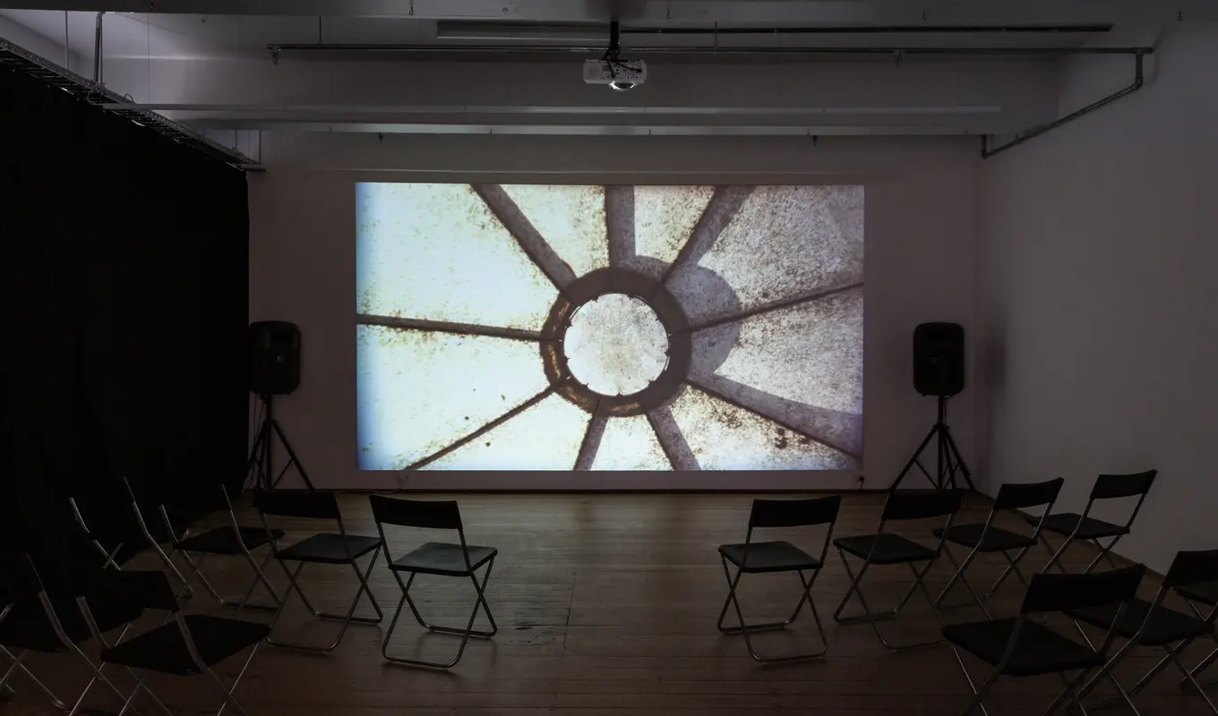 A film is projected on the gallery wall, with two speakers at either side of the image and chairs placed in front. The film still shows