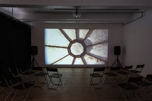 A film is projected on the gallery wall, with two speakers at either side of the image and chairs placed in front. The film still shows