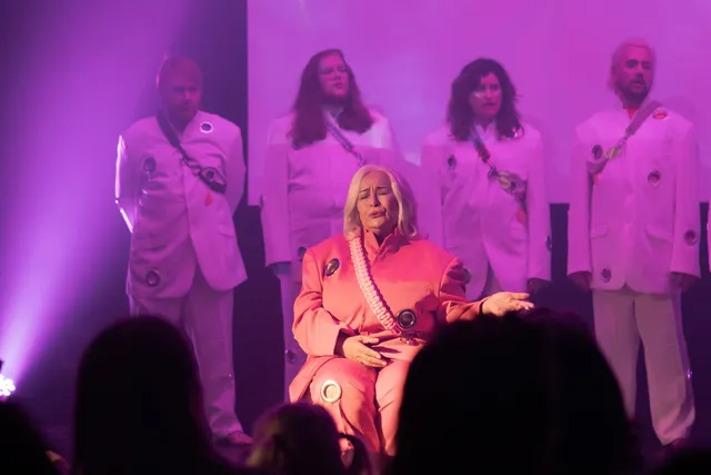 A woman in an orange power suit sits in front of an audience with 4 figures behind her wearing white suits. They are perfoming a musical