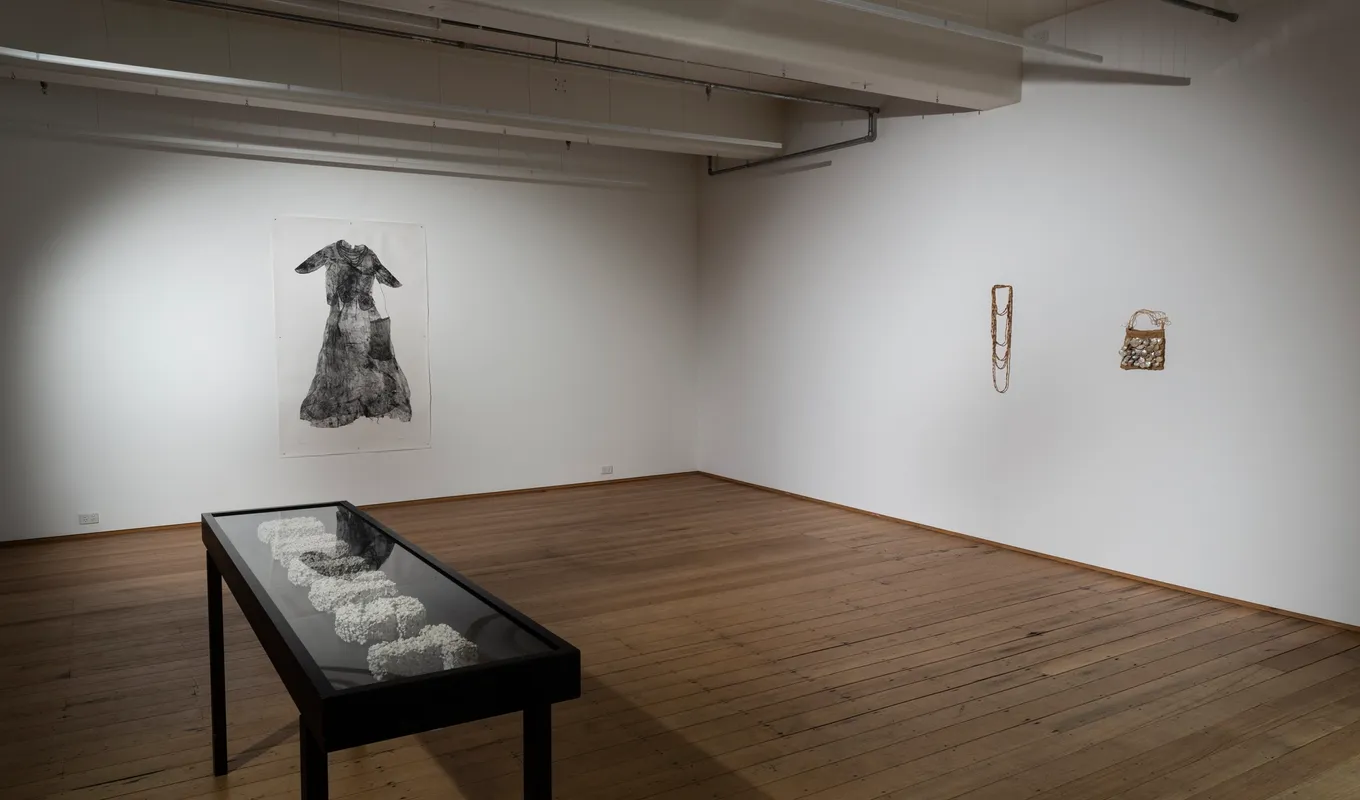 A waist-height cabinet with glass, showcasing a chunky word beneath. On the wall behind is is a large printed dress impression, and on the other wall a shell-covered woven bag and necklace.
