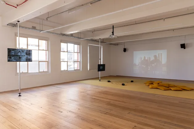 Installation View of 13 Years showing 2 screens suspended away from the gallery wall, one screen has 3 pairs of headphones and some cushions in front of it. The back wall shows a projection of archival footage of two people sitting on a fence in front of 3 large apartment blocks.