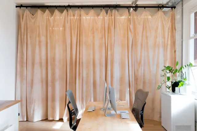 A dyed transparent curtain hangs in the West Space office. It is gathered at the top, creating fluid, overlapping folds and pleats, and the organic dyes create a flush of warm, natural hues. The office is furnished with a desk, chairs, computers and indoor plants.