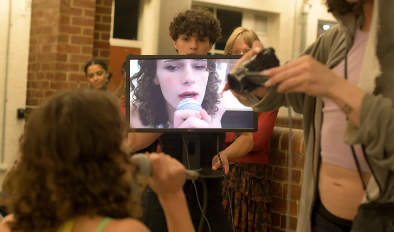 People watching a person speaking to the microphone in front of a screen, footage displaying real time by the other person filming her with a camcorder.