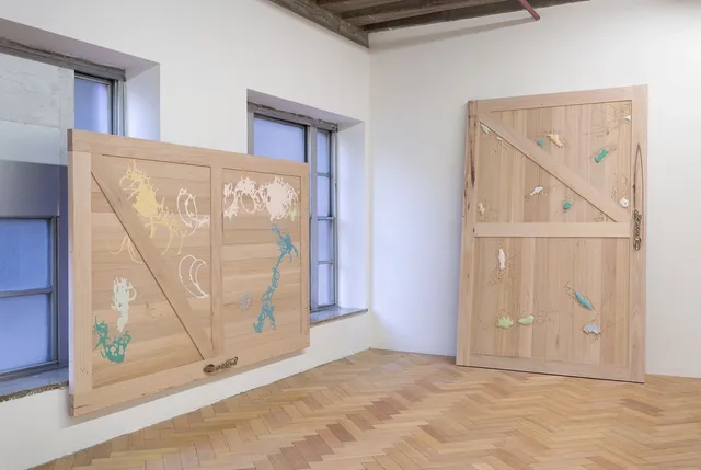 Two of Fiona Abicare's barn door sculptures situated in the corner of the gallery against the wall and hanging on the wall with windows. They each have a decorative brass handle.The doors is adorned with blue, white and yellow and pink painterly and abstract shapes. squiggly lines are carved into the wooden surface around the colourful shapes.