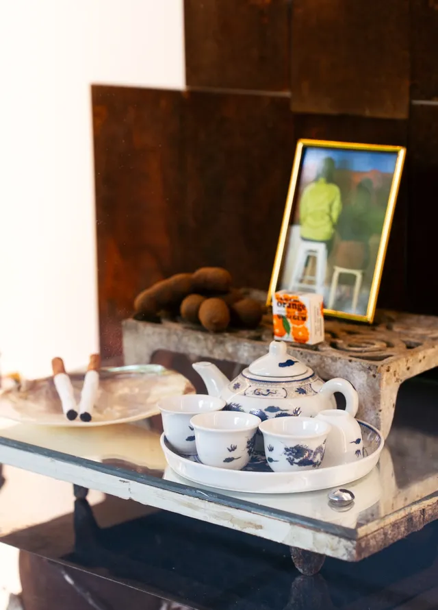 A small teacup set on a ceramic tray, two cigarettes on a flat shell, a small box with some orange graphics on it, some small kiwi-fruit like forms and a photograph of two figure's backs sitting on chairs in a frame, rests on a mirrored, elevated platform.