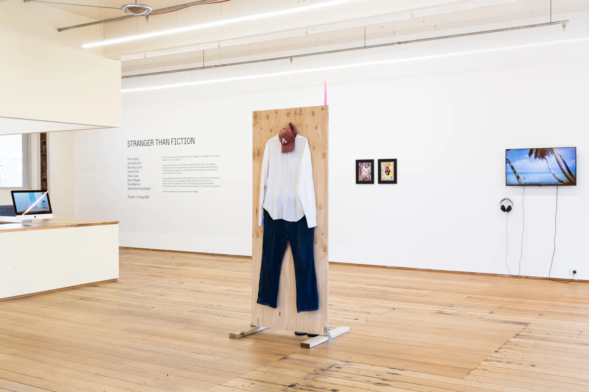 A wide angle installation shot of West Space gallery. From left to right: exhibition text on the wall with the heading "Stranger than fiction", a free standing large piece of ply board with a full outfit of white shirt and dark blue pants and red hat is suspended off the board, 2 framed colourful paper works in frames on the wall and a television screen displaying a beach and palm trees.