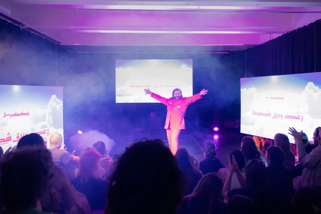 A male figure in a pink suit is posing with jazz hands in front of a small crowd and surrounded by three prominent glowing screens. The space is foggy.
