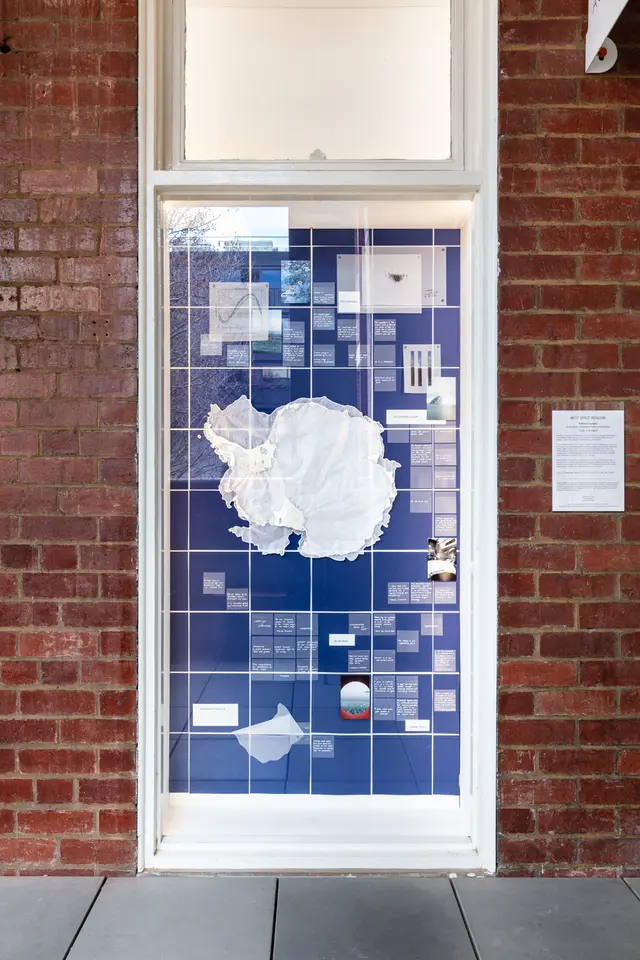 A photo fo the window installation in full. The window shows a background of deep blue squares with white intersections. There are many square notes with white text, photographs, and sheer crinkled white fabric displayed against the background.