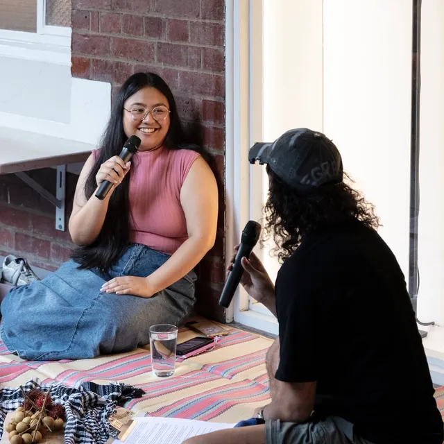 Two figures are in discussion holding microphones at the front of the artists' exhibition window display. The window is exhibiting a screen with text installed on a large black vertical pole. They are both smiling and in dialogue assumably about the work. In front of them is an open blanket-like cloth holding a chopping board covered in a specific plant-like matter or material.