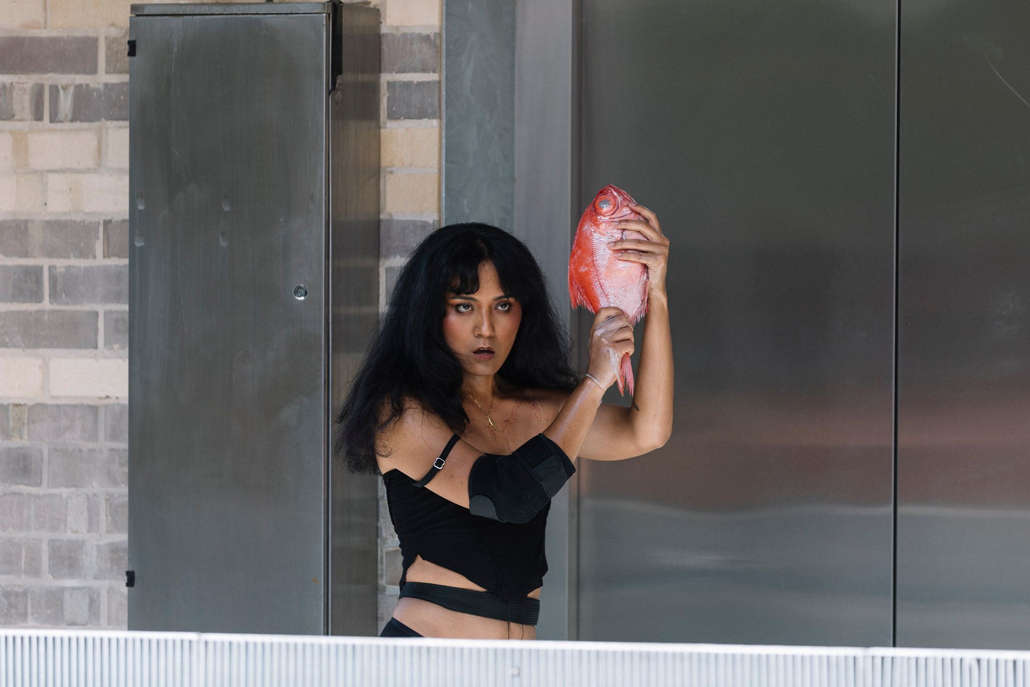 Ari holding a fish like a loaded gun standing in front of silver elevator doors at the Collingwood Yards.