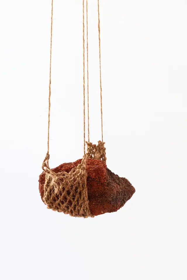 A piece of earth ochre suspended by an open net-like textile made of natural fibres.