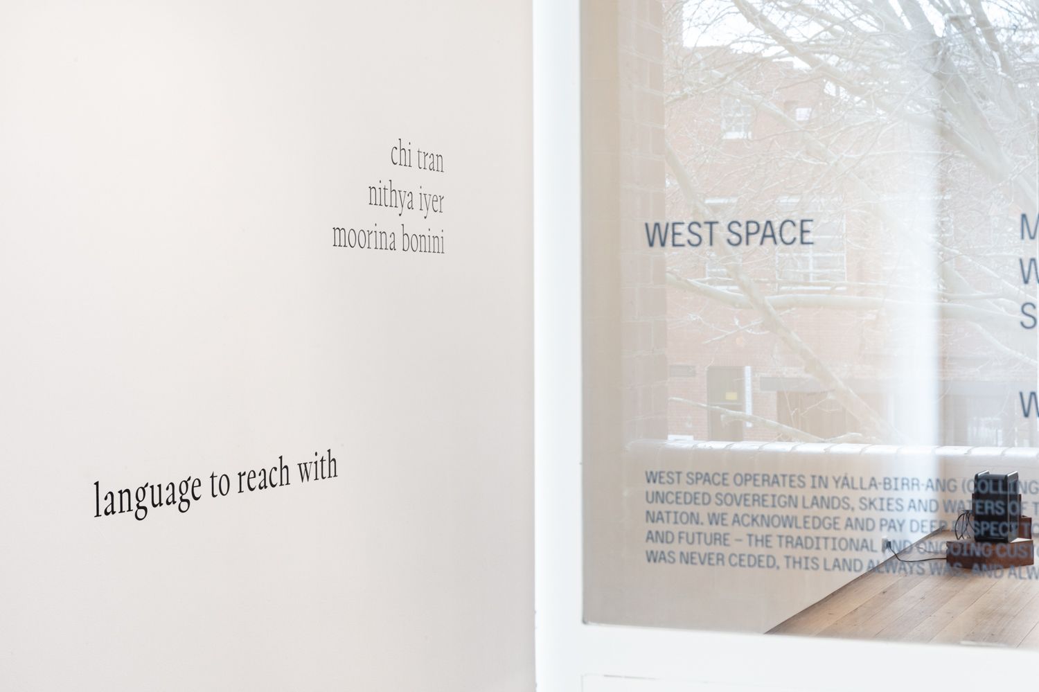 Close-up of West Spaces gallery wall seen through the front glass door featuring the exhibition title 'language to reach with' and three artist names Chi Tran, Nithya Iyer, and Moorina Bonini.