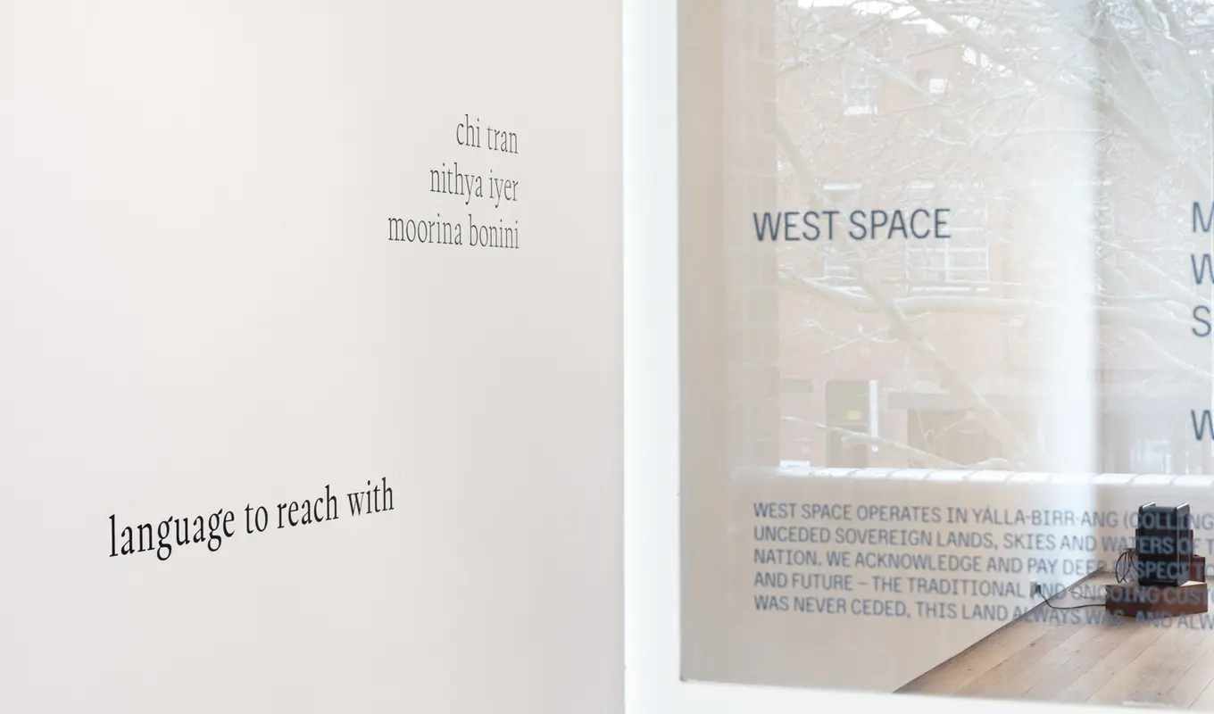 Close-up of West Spaces gallery wall seen through the front glass door featuring the exhibition title 'language to reach with' and three artist names Chi Tran, Nithya Iyer, and Moorina Bonini.