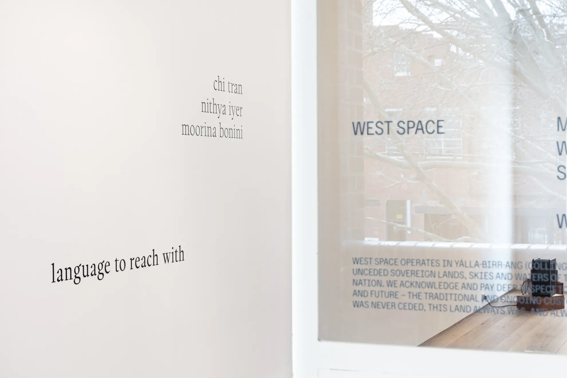 Close-up of West Spaces gallery wall seen through the front glass door featuring the exhibition title 'language to reach with' and three artist names Chi Tran, Nithya Iyer, and Moorina Bonini.
