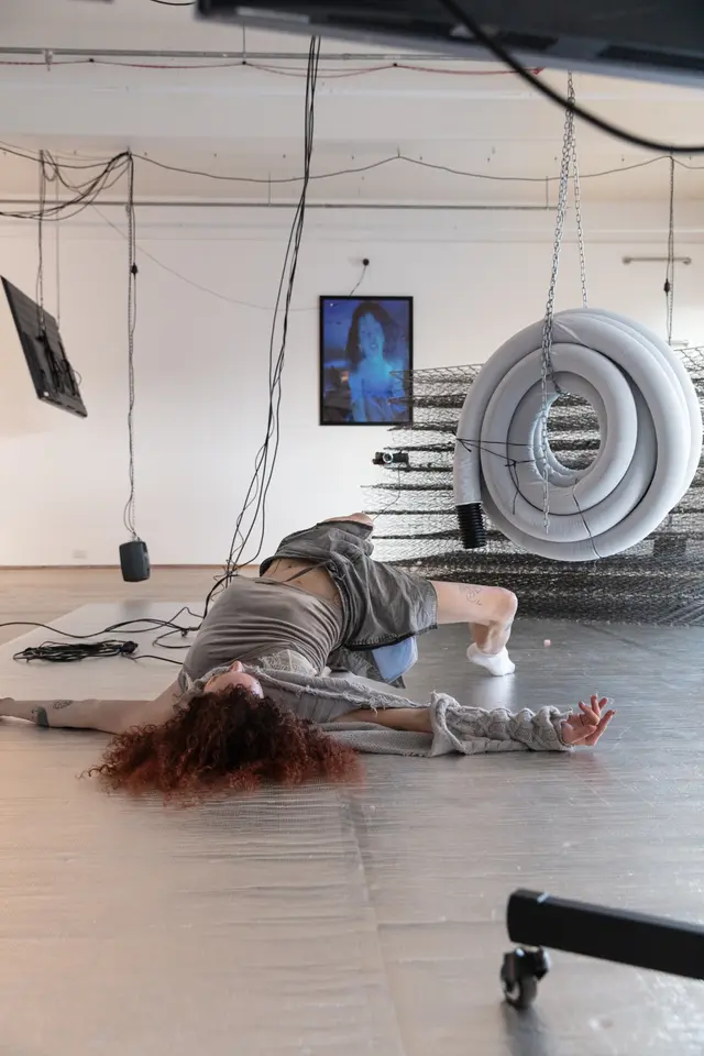 A woman lying on the foil mat on the floor. A coil of plastic pipe that is suspended from the ceiling with chains, a pile of springs from mattresses, and a photo of Andrea are in the background.