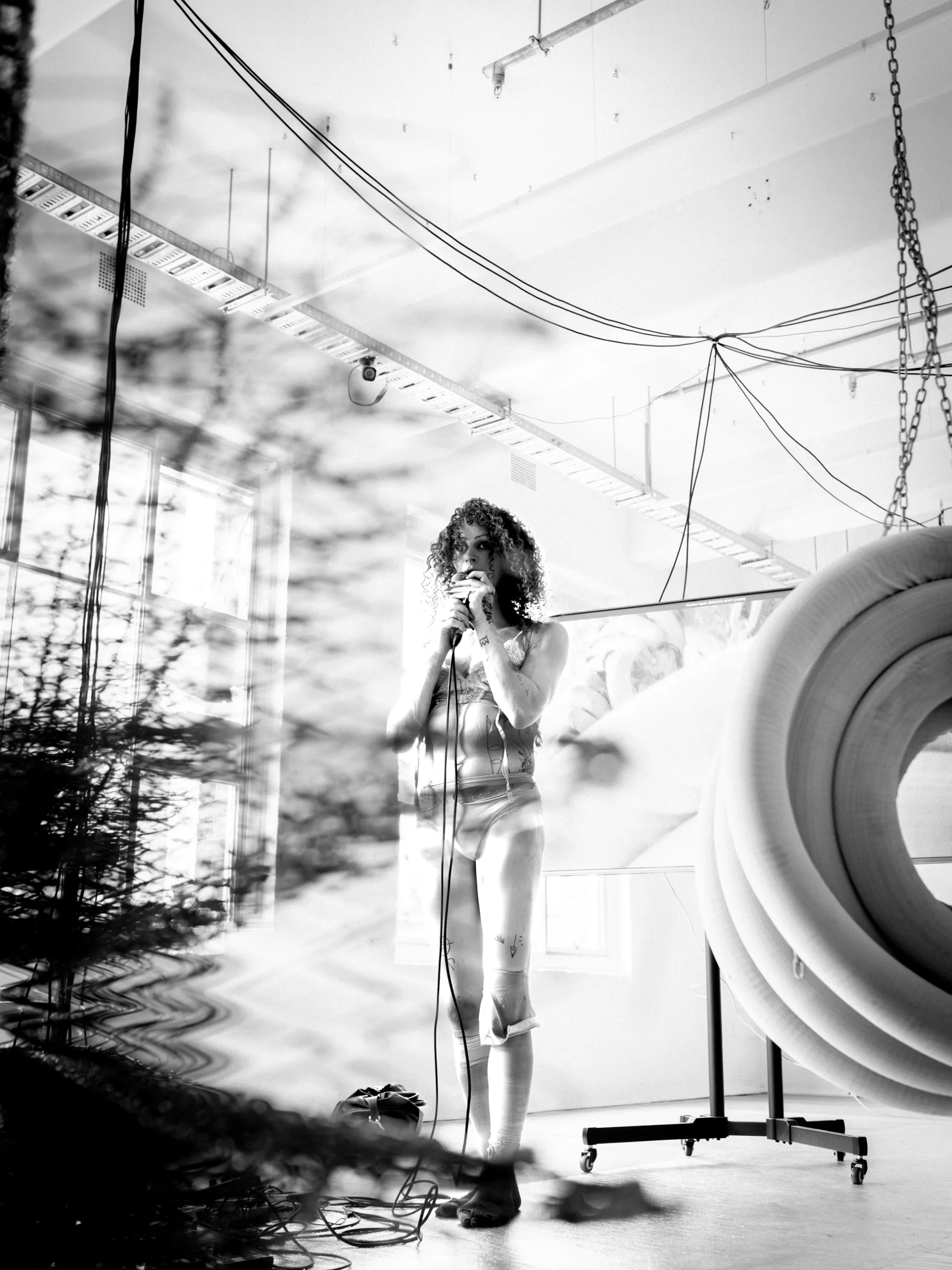Andrea Illés holding up a microphone to her mouth standing amongst tv screens, messy wires and other sculptural elements.