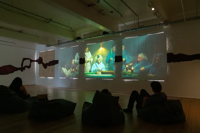People sitting on bean bags in gallery space watching video installation. Three square projections on wall of people sitting and standing in a dimly lit room wearing dressing gowns and weaving at a table.