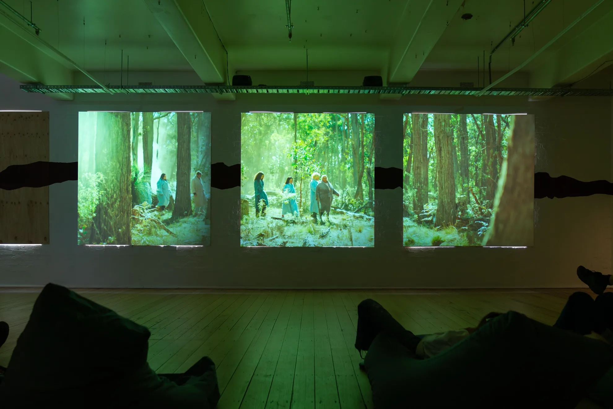 Three square video panels projected of people walking through green bush, with abstract painting in-between. Gallery space with wood floor boards and people sitting on bean bags watching the video.