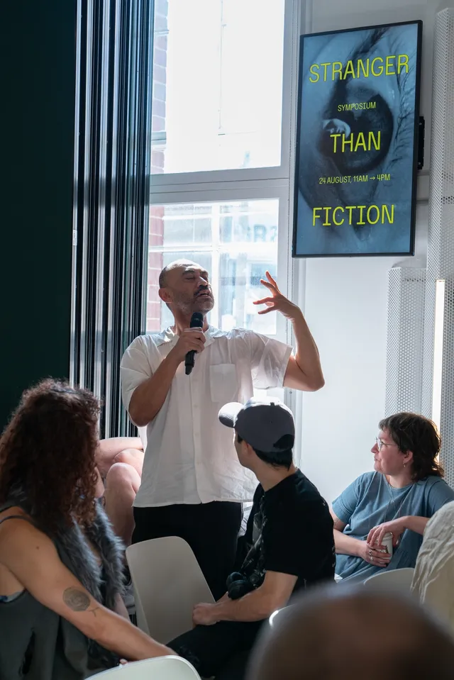 A portrait photo depicts a man in a white shirt performing into the microphone. Around him are three individual crowd members who have their heads turned to watch as he does. There is a screen behind him with an image that reads "Stranger than Fiction", with a photo of an eye behind the text.