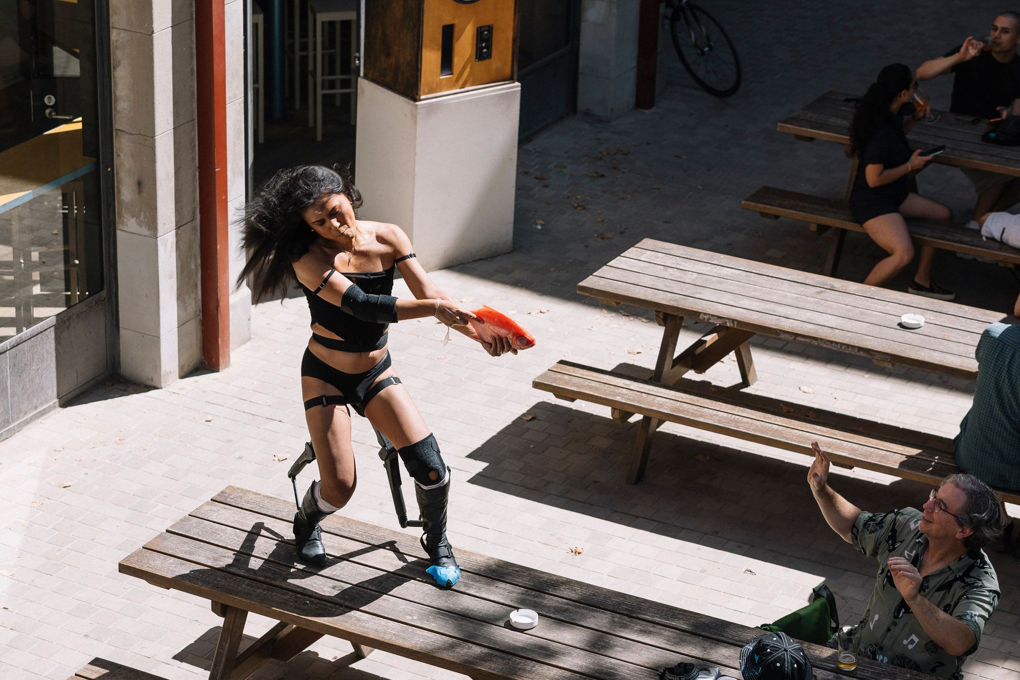 Ari standing on top of a wooden table holding up an orange fish as if it were a gun. In front of her and audience member has their hands up as if to say 'dont' shoot'.