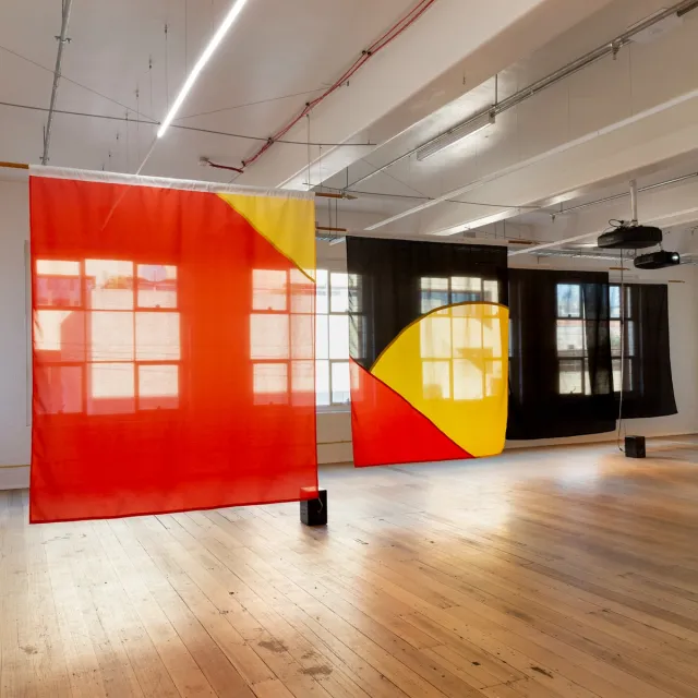 Installation view of "Big Pharmakon". The image shows 4 semi-transparent fabric sheets in front of 3 windows in the West Space Gallery. The first sheet is red with a small triangular yellow section in the top right. The second shows a segment of a yellow circle bisected by a red triangle in the bottom left over a black background. The third and fourth sheets are completely black. On the ground in front of the sheets are two speakers.