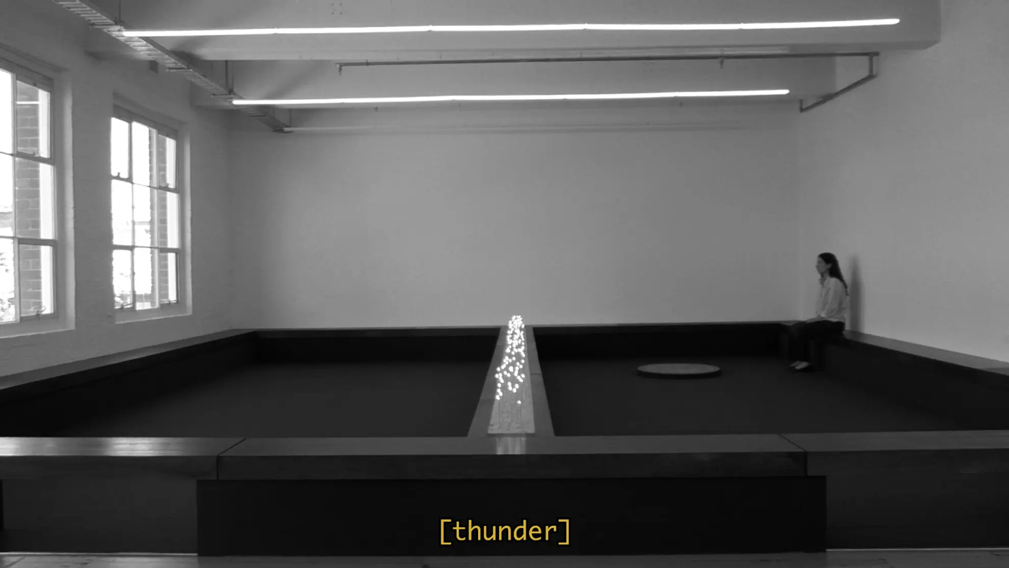 a black and white film still of Tina sitting in the West space gallery install. She is in the far right corner facing the windows and there is a subtitle that reads "thunder".