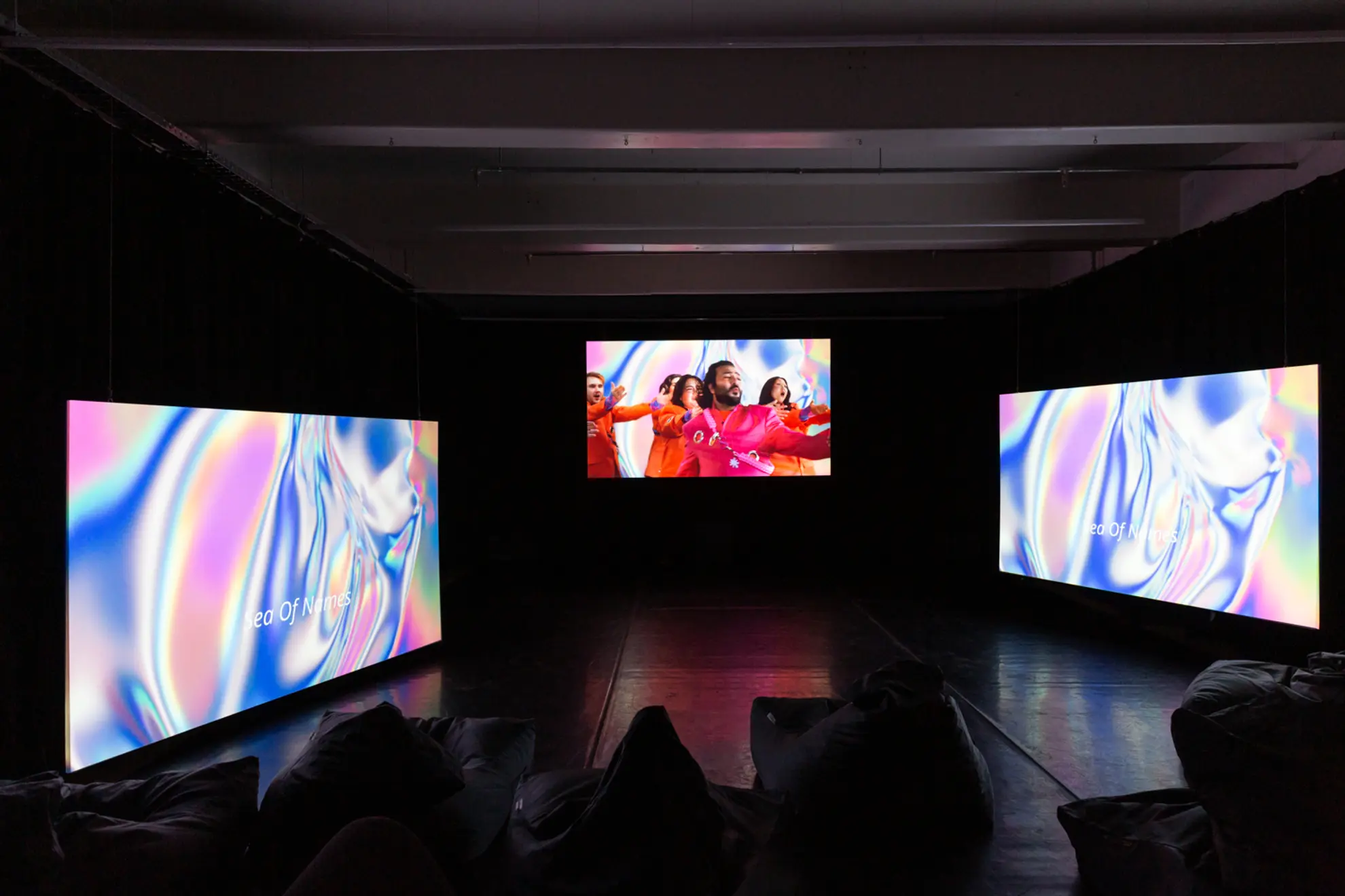 A photograph of artist HOSSEI's audio-visual artwork THUNDERBLOOM. Three screens glow with the projected images of performers, bright backgorund colours and captioned lyrics.