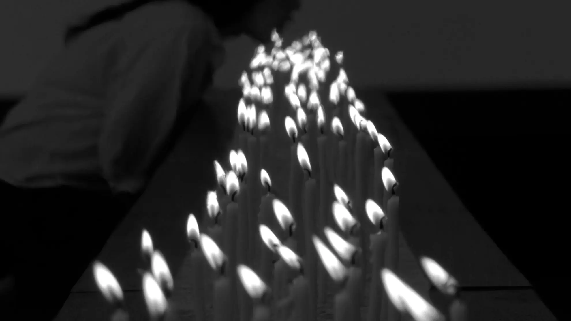 a black and white film still on a long bed of burning candels. In the background there is a shadowed figure on her knees leaning forward to blow on the candles.