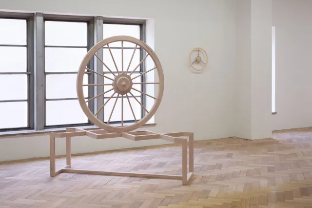 Installation view of a large wooden sculpture or a wheel placed on a bench. The wheel is balancing on the intersection of two pieces of wood crossing over on the bench. The wheel is placed in front of a large window with natural light shinning through. On the wall in the background is a sculpture of a smaller wheel.