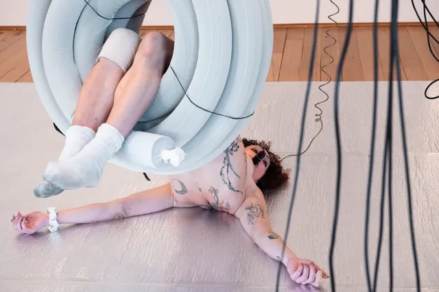 A woman lays with her legs elevated through a suspended coil of pipes. Her arms are straight and sprawled out and she has a camera in her mouth attached to a cord