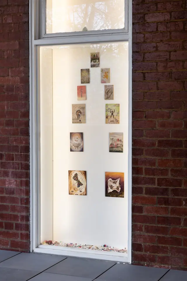 Installation view of Matilda Davis' "I’ll Leave a Secret in the Window for You: Painting Grotto". The installation shows 11 small paintings on the wall and a series of trinkets on the ground all behind a glass panel.
