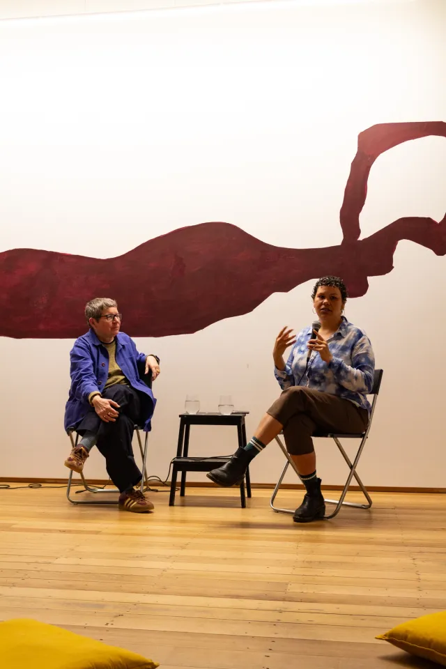 Two people sitting on chairs with a table in-between holding glasses of water, one person speaking into microphone. Maroon abstract painting displayed on the wall behind them and yellow cushions in front of them on the wood floor.