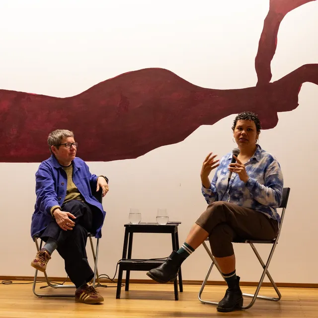 Two people sitting on chairs with a table in-between holding glasses of water, one person speaking into microphone. Maroon abstract painting displayed on the wall behind them and yellow cushions in front of them on the wood floor.
