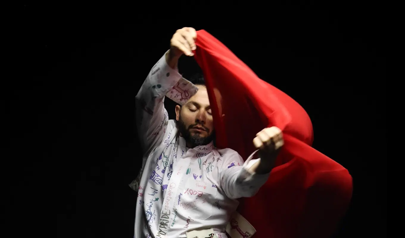 A man wearing a white jump full of drawings is holding a red sheer fabric with both hands.