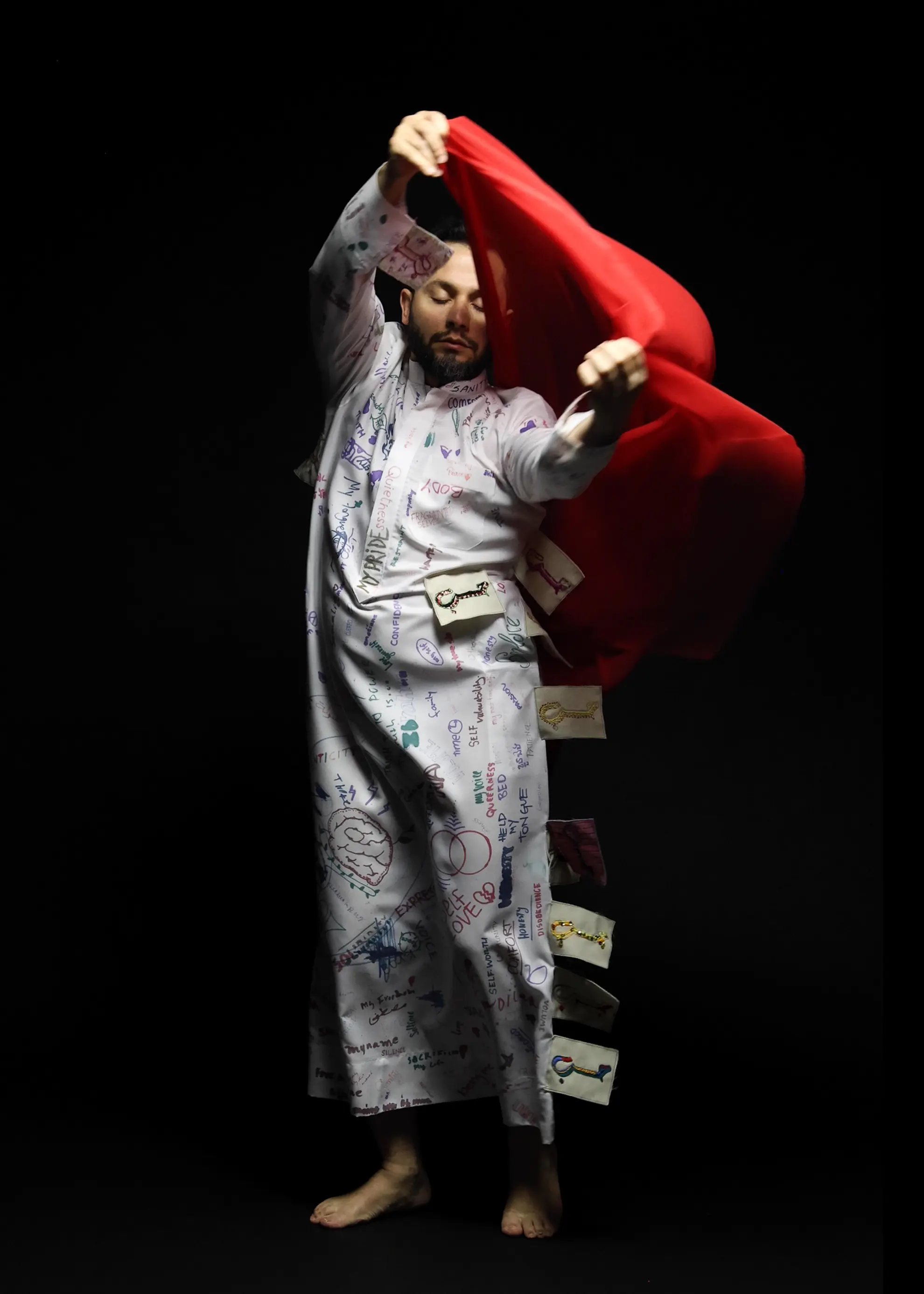 A man wearing a white jump full of drawings is holding a red sheer fabric with both hands.