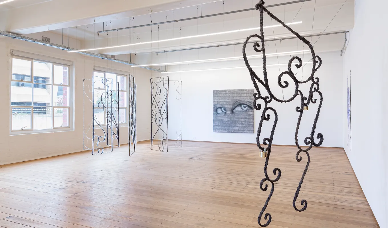 Installation shot of Grace Culley's solo commission show. There is a large textured curly metal sculptural work suspended in the foreground. In the background there is a circle of suspended gate-like artworks and a large painting on the wall with eyes and heart-shaped pupils.