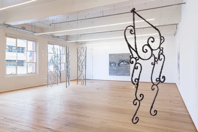 Installation shot of Grace Culley's solo commission show. There is a large textured curly metal sculptural work suspended in the foreground. In the background there is a circle of suspended gate-like artworks and a large painting on the wall with eyes and heart-shaped pupils.