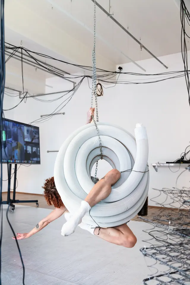 A woman is suspended from the roof hanging in a coiled pipe. One of her legs is in the middle of the pipe.