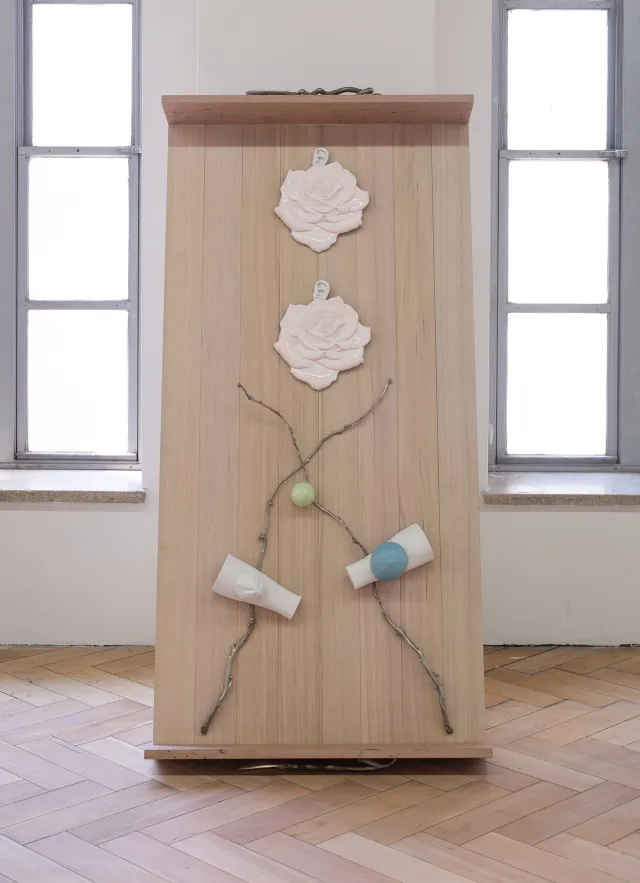 Front view of 'Drawer 1' sculpture in Rose Moon'. The draw is leaning against the wall and light from widows either side of the work is shinning softly through. There are two rose shaped earrings, two brass branches and three other objects in white, blue and green attached to the wooden draw. Two brass handles are attached to the sculpture on the top and bottom.