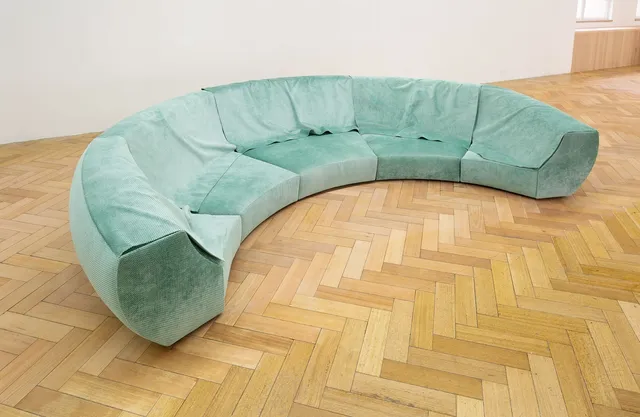 A sculpture of a five seater half moon shaped lounge is situated  on the floor of the gallery. The fabric is a light teal colour that appears to be soft the touch. There is excess fabric on the work that is draped over the back of the lounge to form a flap or cover.