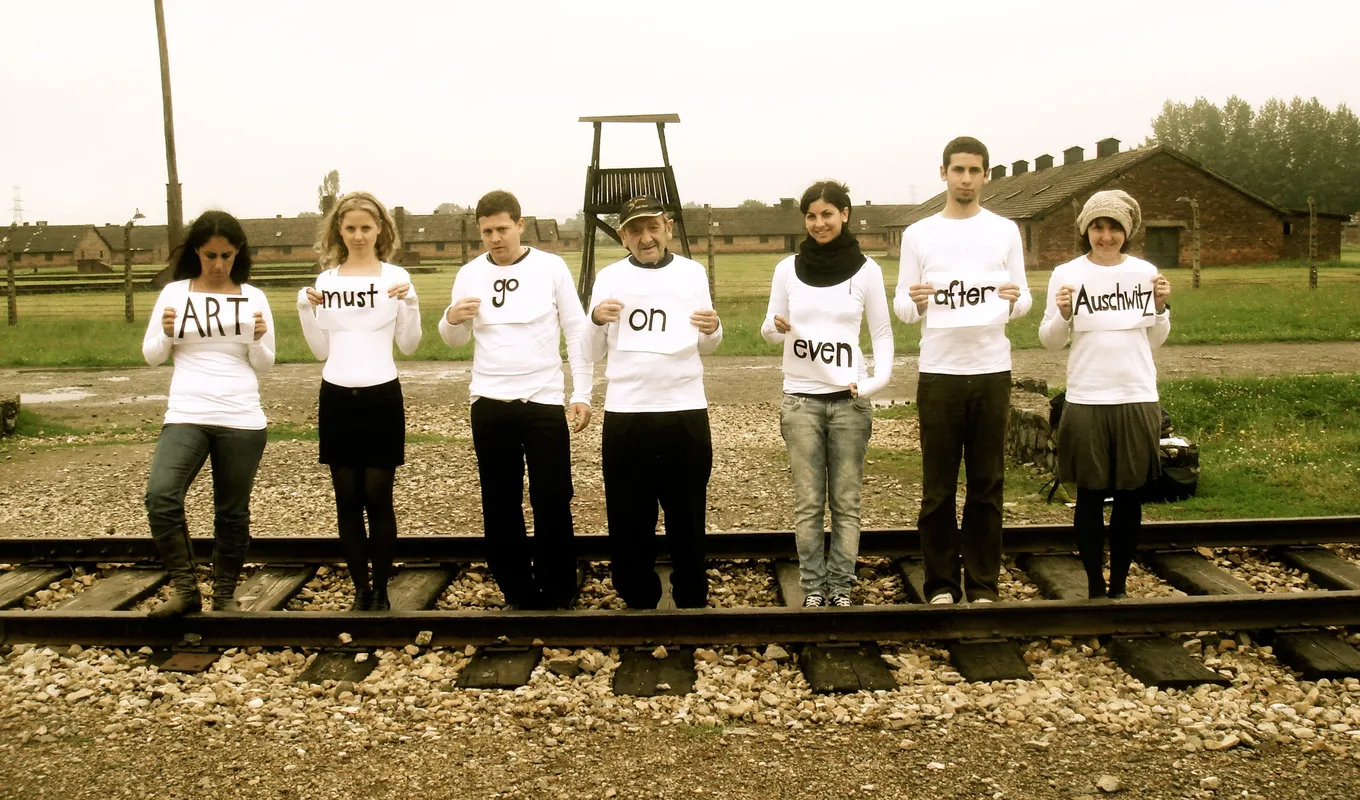 A row of people standing on a train track each holding a paper sign. From left to right they read 'Art must go on even after Auschwitz'.