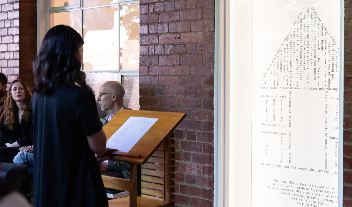 A figure wearing black reads text from a wooden platform whilst facing a window display. Behind the tall window pane is a tall house embodying and silhouetted by text, perhaps in relation to what the individual is reading. A couple figures in the backdrop listen intently.