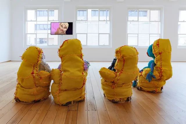 Installation view of Terry Williams' soft sculptures. The yellow fabric back of the sculptures are visible with orange and red seams showing where the fabric has been sewn together. The sculptures are faced towards Victoria Todorov's video work, appearing as though the sculptures are watching it.