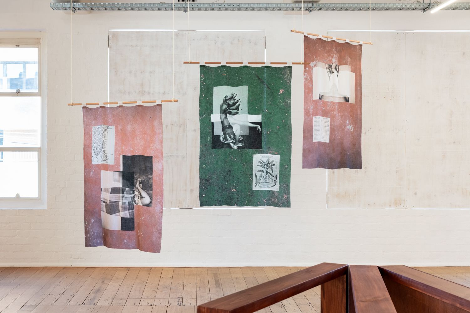 Three printed textile works hang on wooden poles from the West Space gallery ceiling the two on the left and right are a dusty pink and the one in the middle is a deep green. The windows behind this work have been blocked out by white boards.