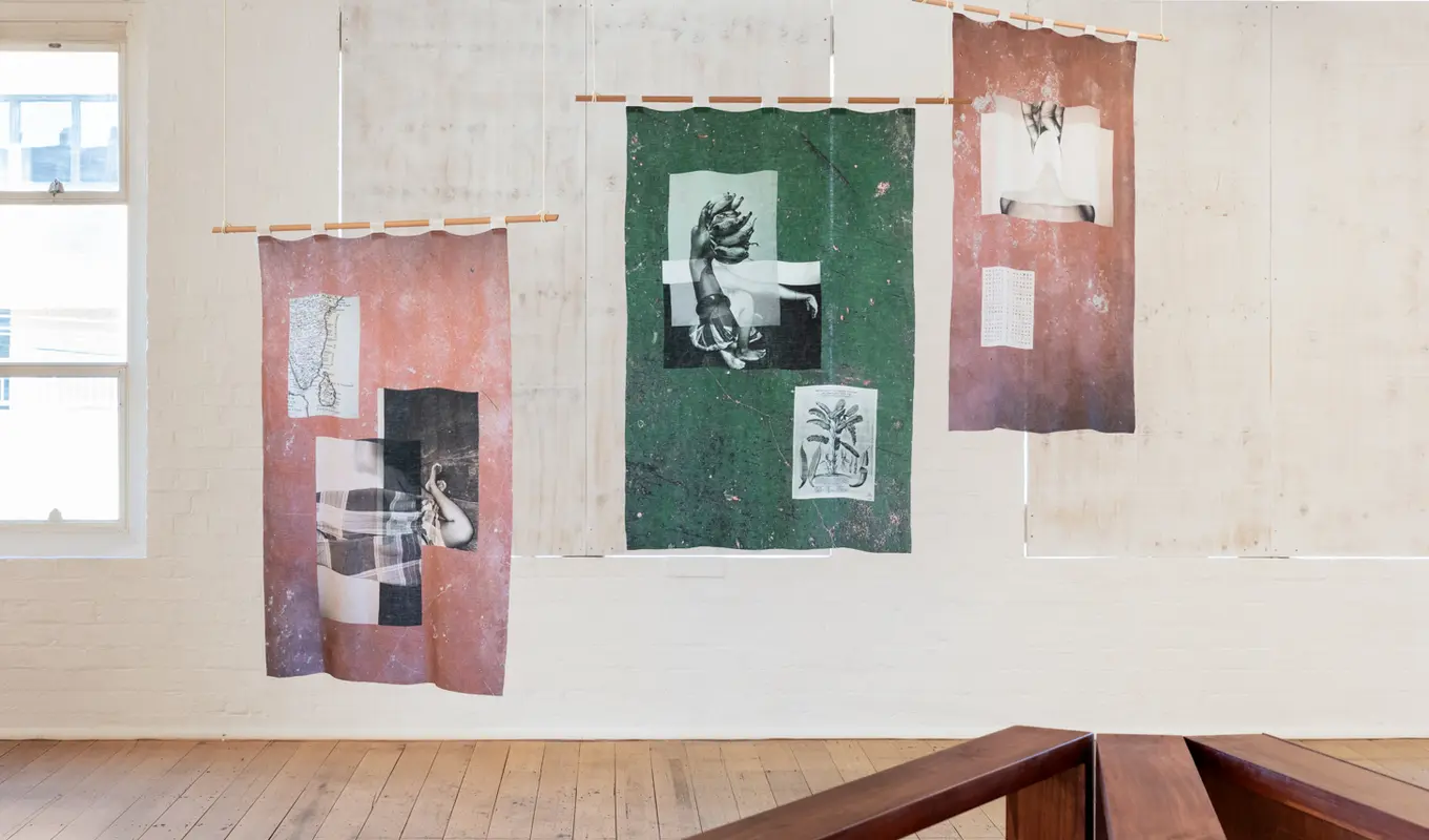 Three printed textile works hang on wooden poles from the West Space gallery ceiling the two on the left and right are a dusty pink and the one in the middle is a deep green. The windows behind this work have been blocked out by white boards.