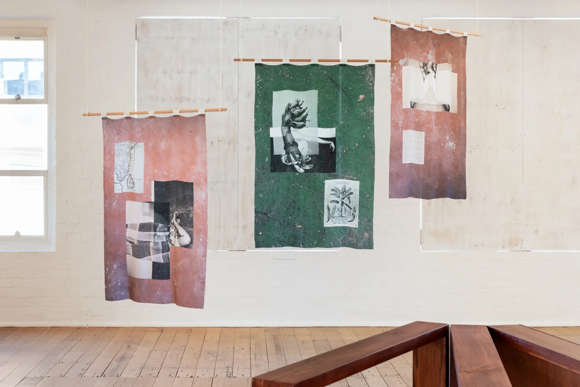 Three printed textile works hang on wooden poles from the West Space gallery ceiling the two on the left and right are a dusty pink and the one in the middle is a deep green. The windows behind this work have been blocked out by white boards.