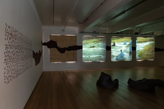 In a dark room, there are 3 large squared transparent images of some figures in a green filend. The room is neatly lined with been bags for viewing.