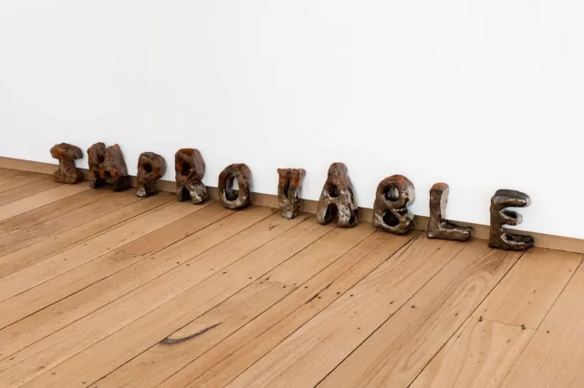 Ten small hand crafted metal letters sit on the floorboards, leaning against a white wall. They spell out 'IMPROVABLE'.